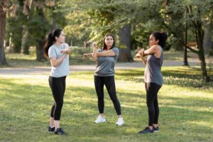 Three women stand on grass in a park, facing each other and performing arm stretches, dressed in athletic wear. Trees and greenery are visible in the background.