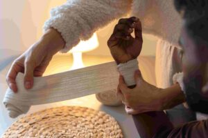 A person wraps a bandage around another person's wrist, with a woven placemat and a lamp in the background.
